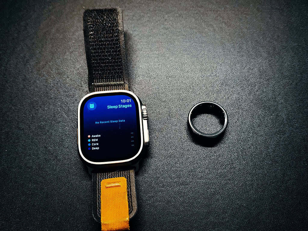 An overhead shot of a silver smartwatch with a black and orange strap, displaying its "Sleep Stages" screen which reads "No Recent Sleep Data." Next to it lies a black, matte-finish smart ring on a dark, textured surface.