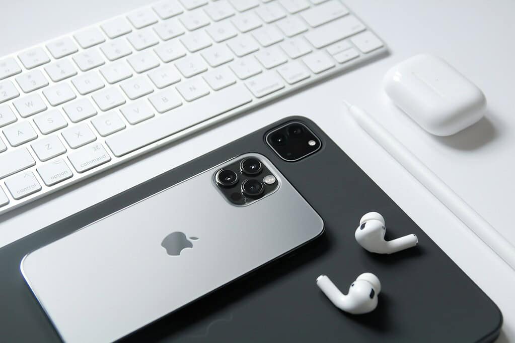 Un élégant iPhone et iPad Apple sont à côté d’AirPods blancs et d’un clavier, tous disposés sur une surface de bureau minimaliste.