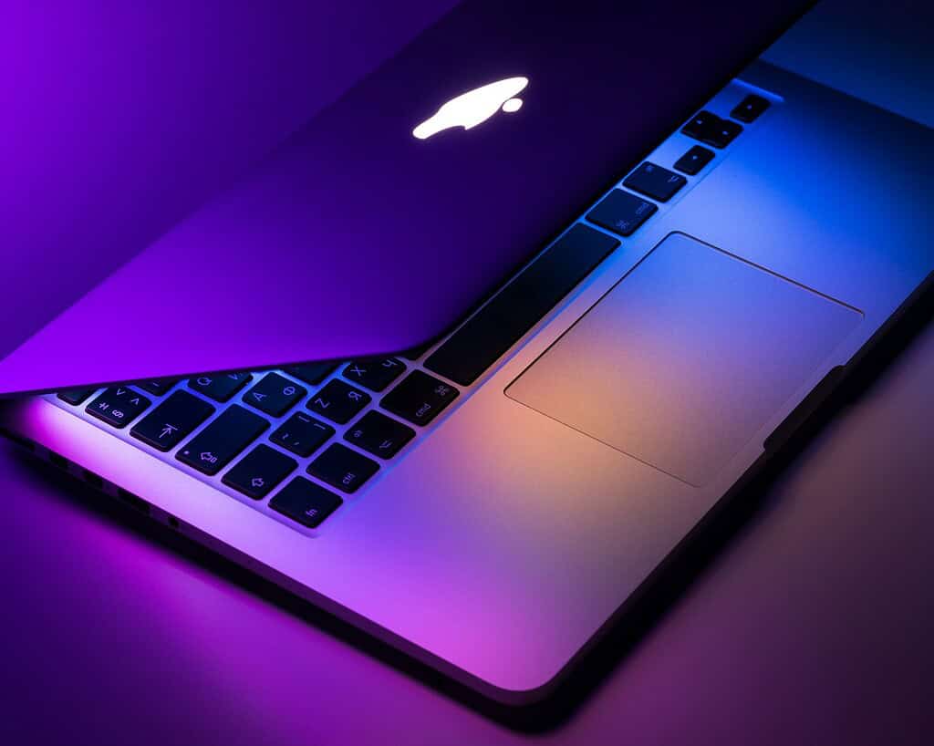 Een close-up van een gedeeltelijk geopende MacBook, verlicht in levendige paarse en blauwe tinten, die het strakke toetsenbord en trackpad toont.