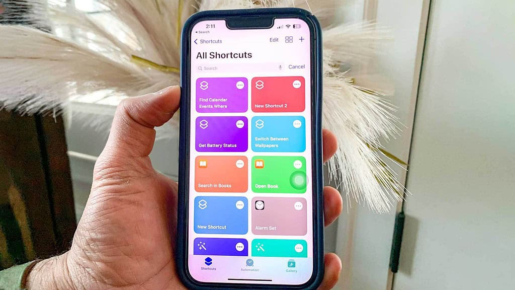 A hand holds a smartphone displaying the "Shortcuts" app with various colorful icons for automation tasks like "Get Battery Status."