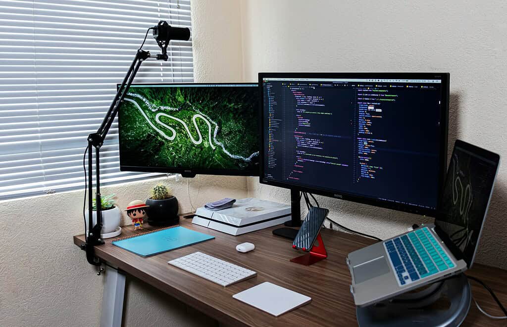 Un espace de travail moderne avec deux écrans, du code sur l’un, un ordinateur portable et des plantes en pot, avec une vue extérieure dynamique à travers des stores.