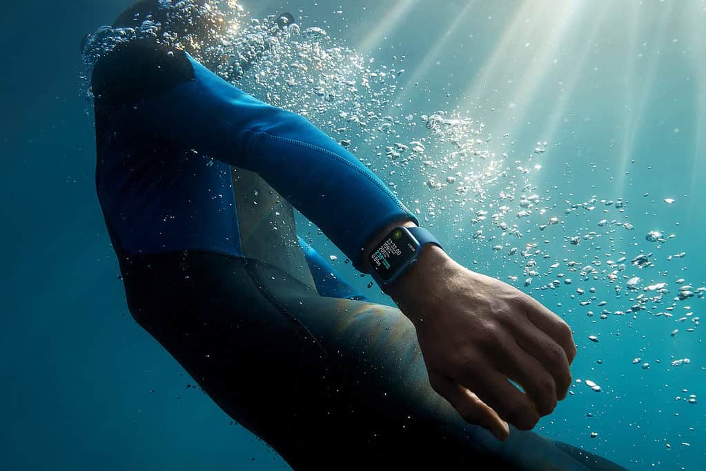 A swimmer underwater checking an Apple Watch with dive metrics, surrounded by bubbles and sun rays.