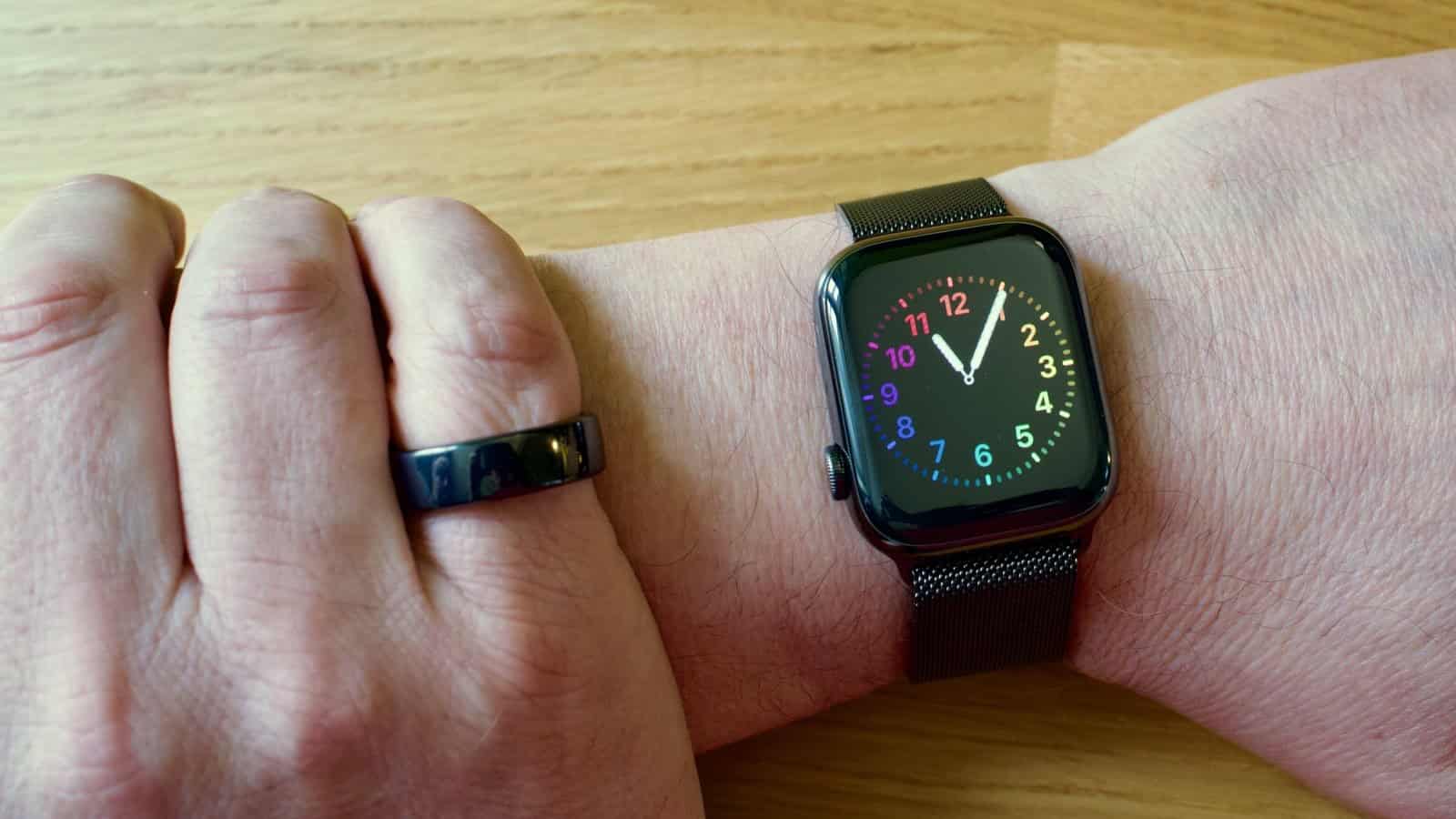 A close-up of a person's wrist wearing a black smartwatch with a rainbow-colored analog clock face. The watch has a mesh band and is paired with a sleek, black ring on the index finger. The background is a light wood surface.