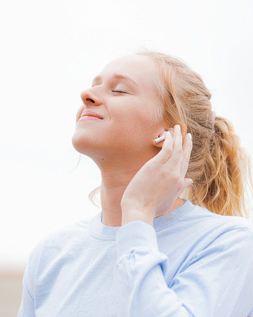 Una persona con una camisa de manga larga azul claro está de pie al aire libre con un cielo brillante de fondo, sosteniendo los AirPods en su oreja, participando en una conversación.