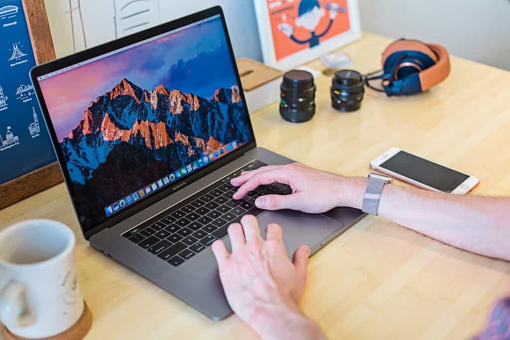 Een persoon die typt op een MacBook Pro, omringd door een koffiemok, smartphone, cameralensen en koptelefoon op een houten bureau.