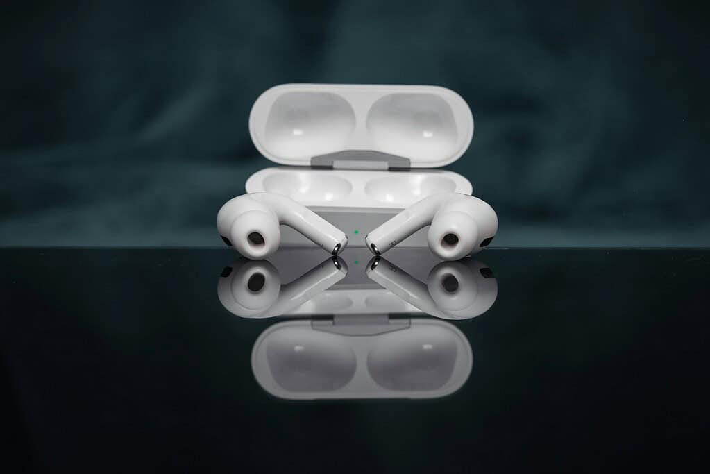 Dos auriculares inalámbricos blancos yacen junto a su estuche de carga sobre una superficie reflectante, con un fondo oscuro suave.