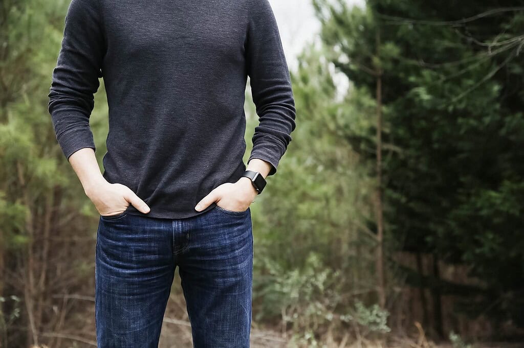 A man standing outdoors in casual clothing, wearing a dark long-sleeve shirt and jeans, with a smartwatch on his wrist and hands in his pockets.
