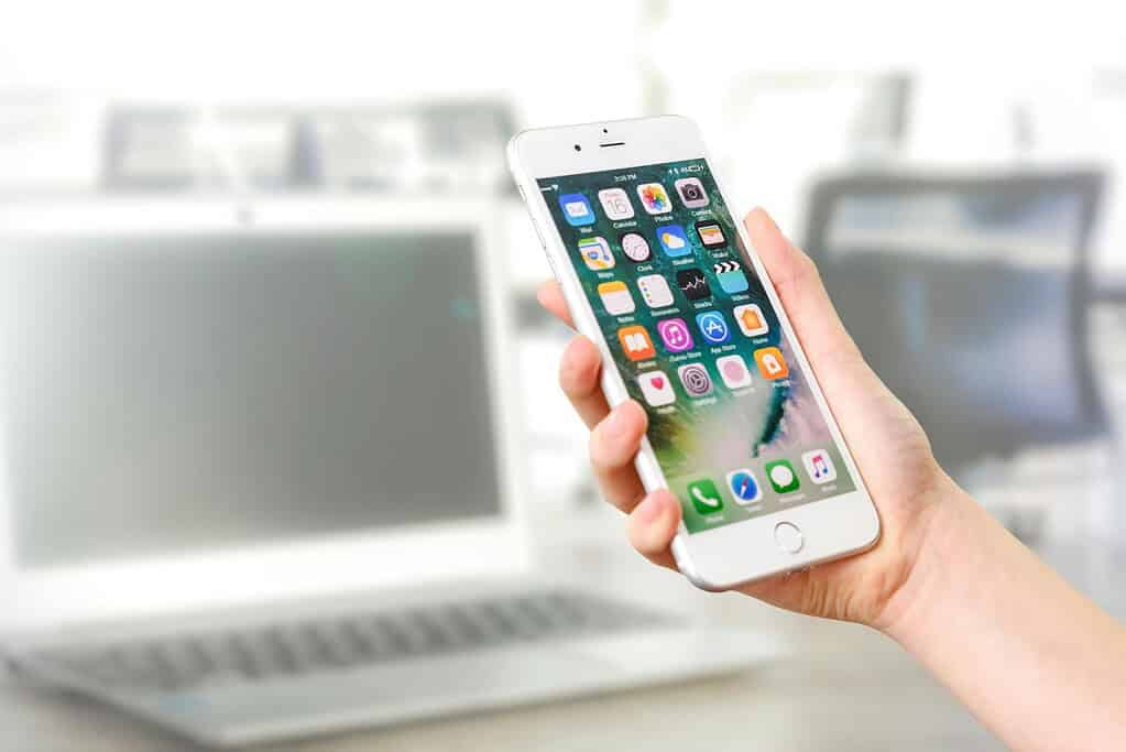 Una mano sostiene un iPhone que muestra varios iconos de aplicaciones, con un portátil borroso en el fondo.