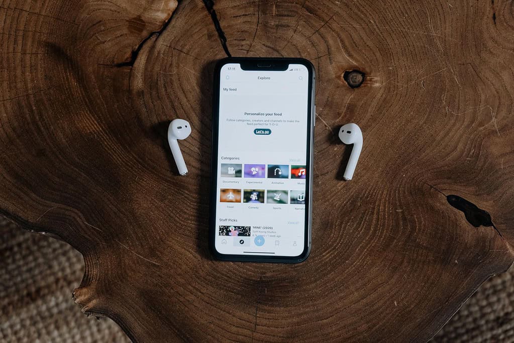 A smartphone displaying an "Explore" feed on a wood table, flanked by two Apple AirPods. 