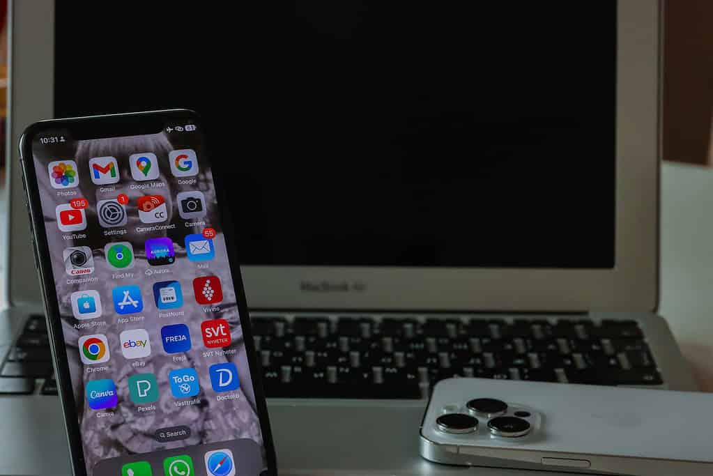 A smartphone displaying apps in front of a MacBook Air, with a silver iPhone partially visible on the right.