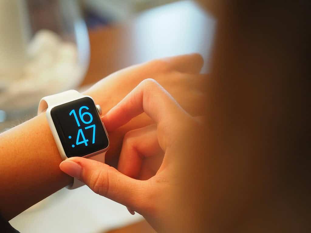 A close-up of a hand adjusting a white smartwatch displaying the time as 16:47, with a blurred background.