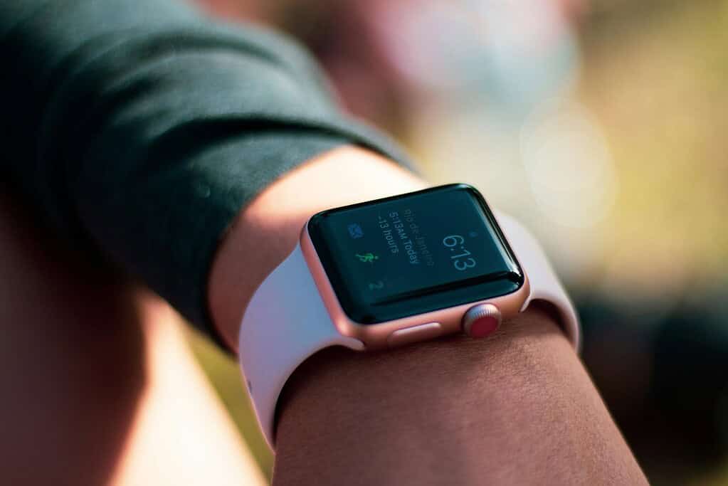 A close-up of a wrist wearing an Apple Watch displaying the time 6:13 and weather details for Rio de Janeiro on a sunny background.
