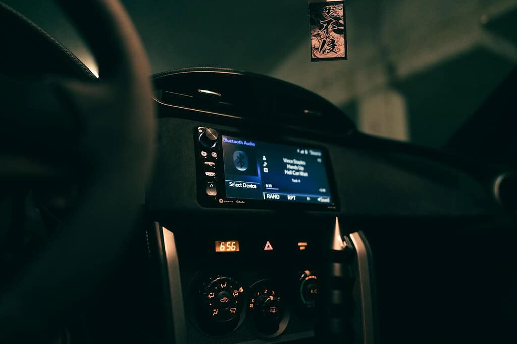 Close-up of a car's dashboard featuring a Bluetooth audio screen, climate controls, and a hanging ornament in dim lighting.