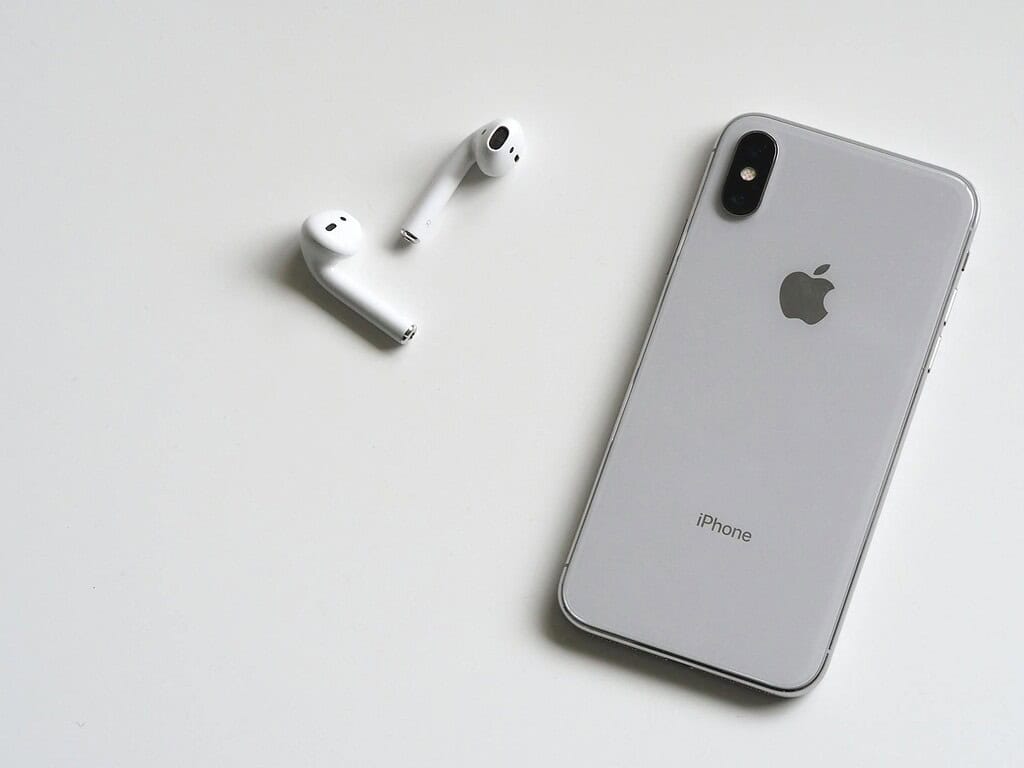 An iPhone with a silver back lies on a white surface next to a pair of white Apple AirPods.
