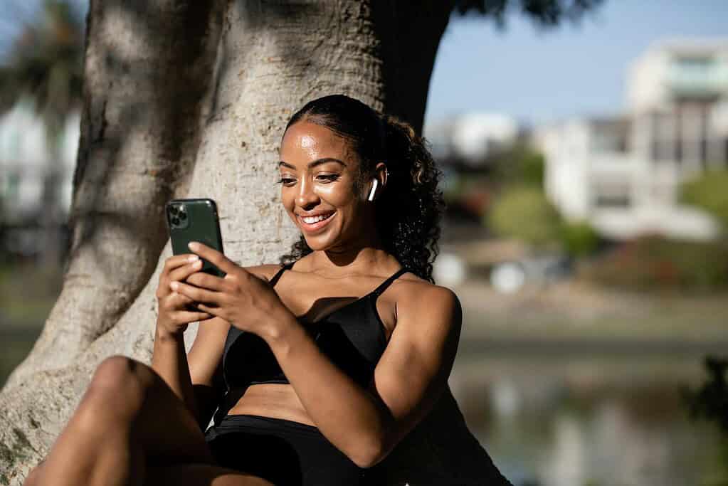 A person sitting under a tree, wearing a black bikini top and earbuds, focused on their smartphone by a serene water scene.