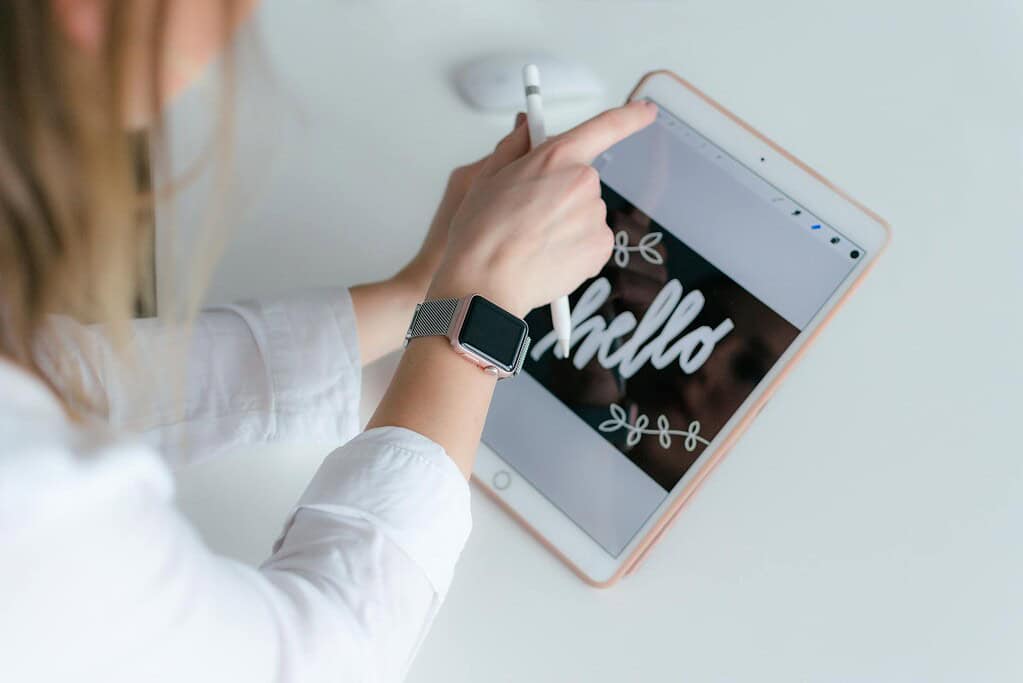 A person writes "hello" with a stylus on a tablet, wearing a silver mesh watch and a white shirt.