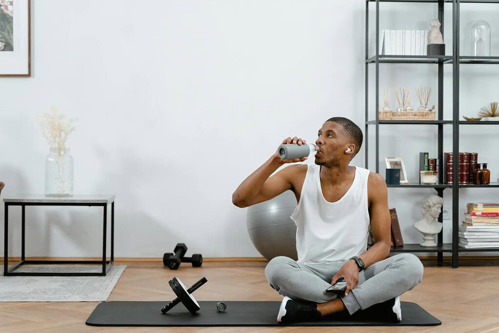 A young man sitting on an exercise mat in a modern home gym, drinking from a bottle, with workout equipment nearby.