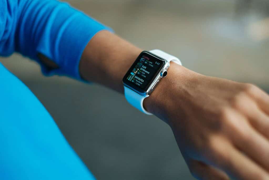 A close-up of a hand wearing a white smartwatch displaying nearby cafes and their distances, against a blurred background.