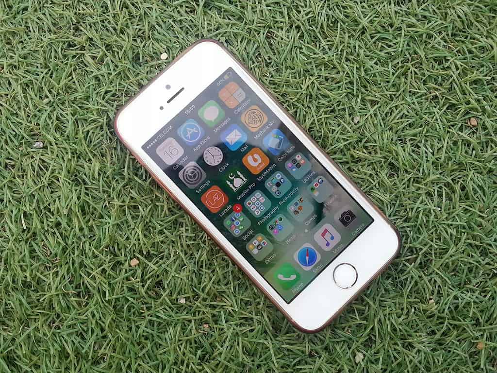 A white iPhone 5S lies on green artificial grass, displaying its home screen filled with various app icons.
