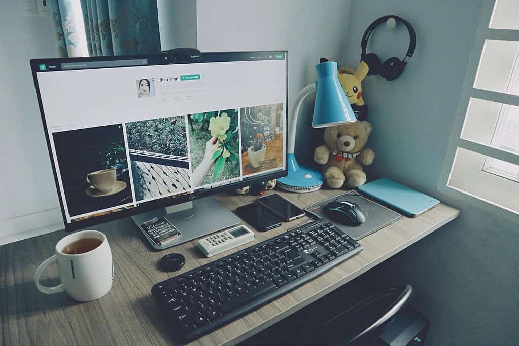 Une configuration de bureau comprenant un écran d’ordinateur affichant des images, un clavier, une tasse de thé et des objets décoratifs comme une lampe bleue et un ours en peluche.