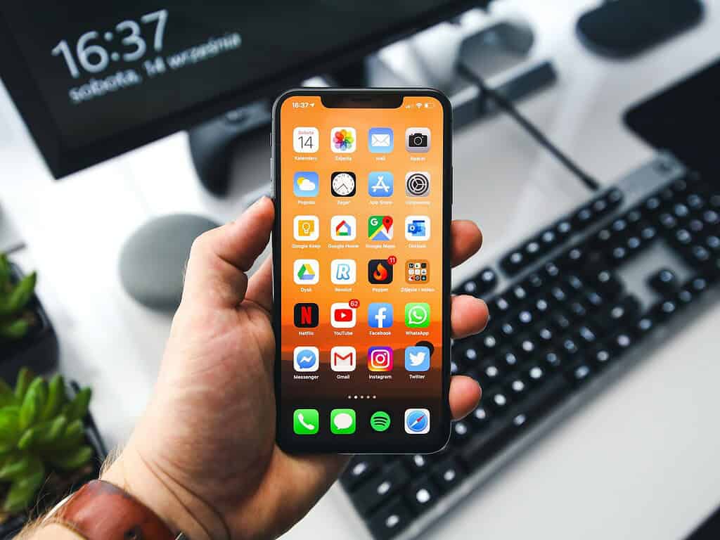 Iemand houdt een smartphone vast die een beginscherm vol apps toont, met een vervaagd toetsenbord en monitor op de achtergrond.