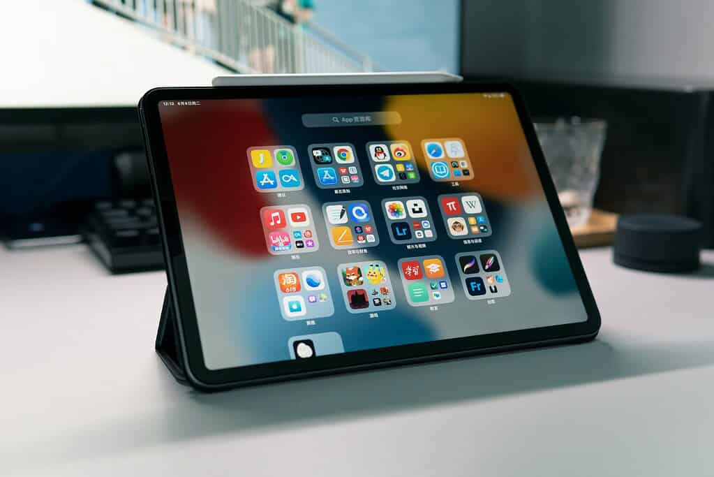 A tablet on a desk displaying various app icons, with a blurred background of a monitor and other workspace items.