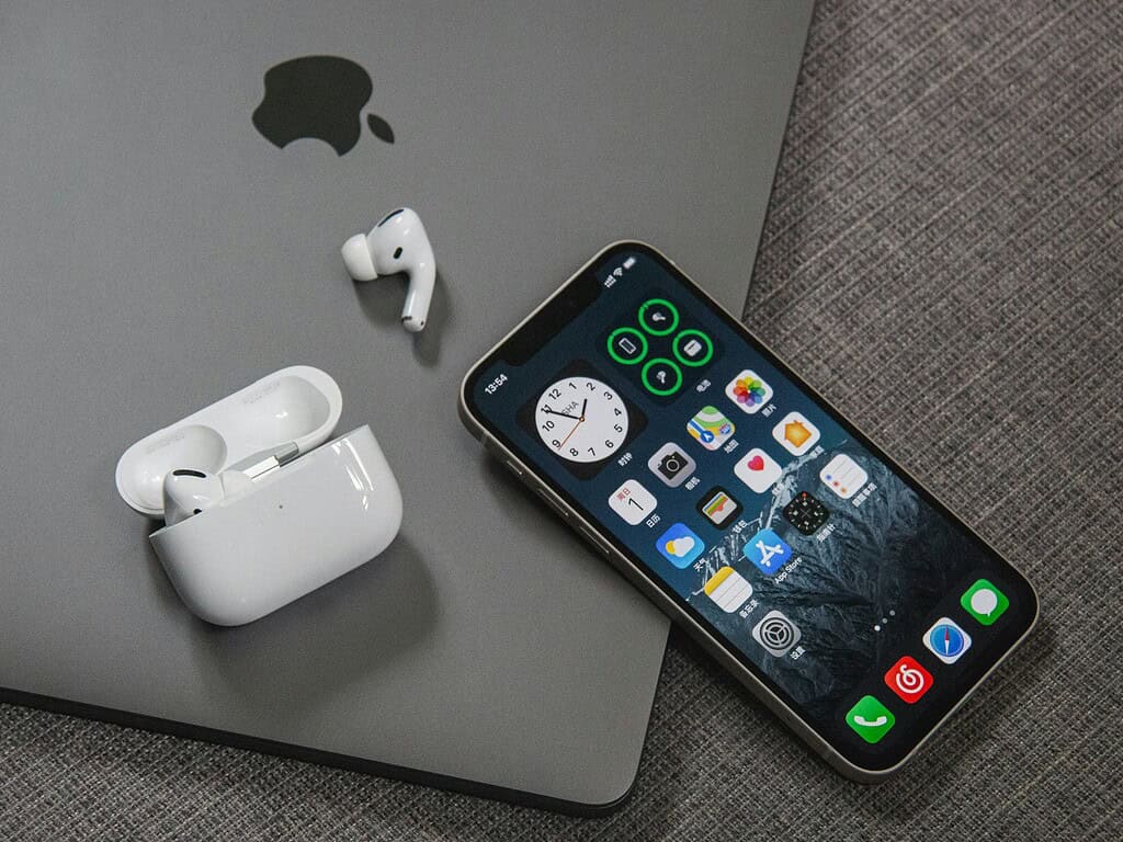 A pair of AirPods is next to their charging case, beside an iPhone displaying a colorful app grid and a clock, on a gray laptop.