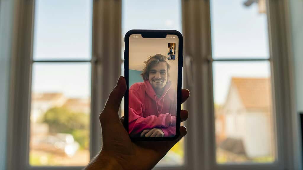 A person video chatting on a smartphone, holding it in one hand, with a bright background of windows and rooftops.