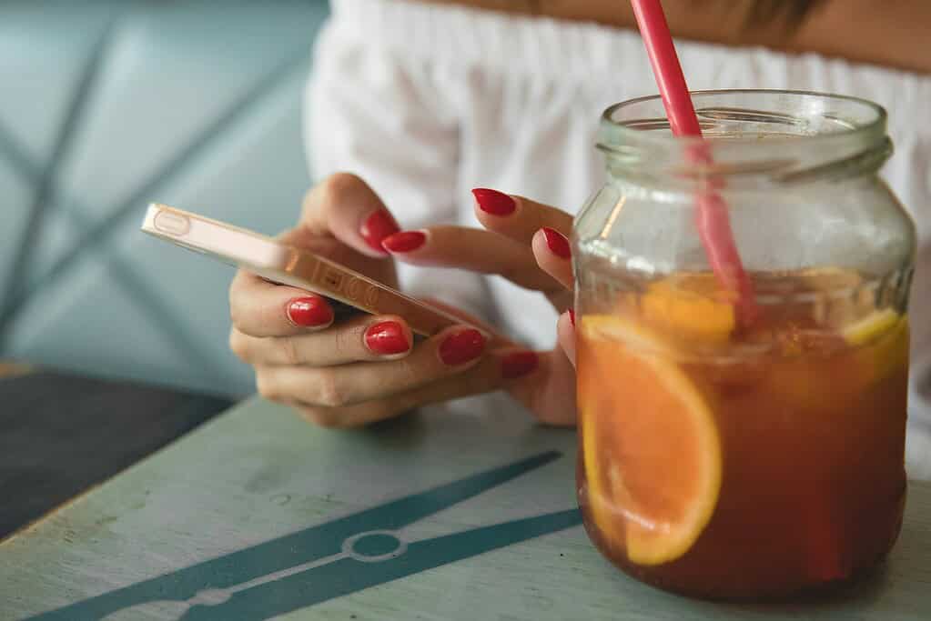 A person with red nails texting on a smartphone next to a jar drink filled with orange slices and a pink straw.