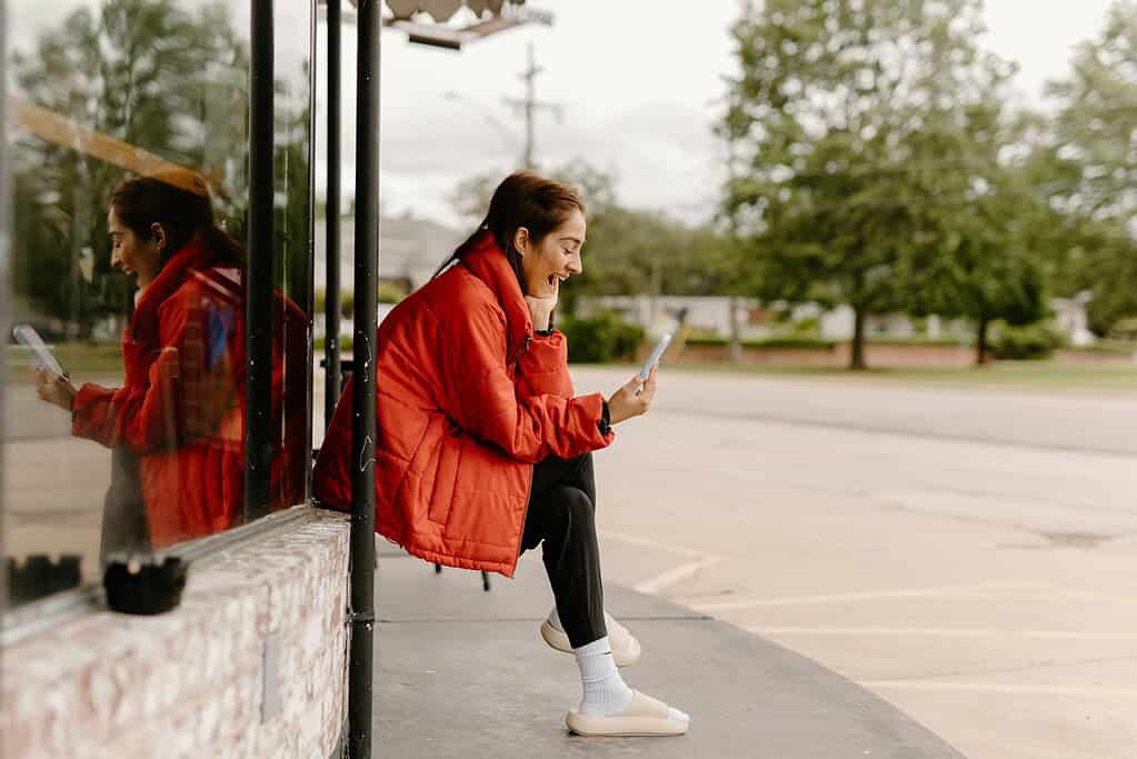 A person in a bright red jacket sits outside, intently looking at their phone, reflecting in the glass window beside them.