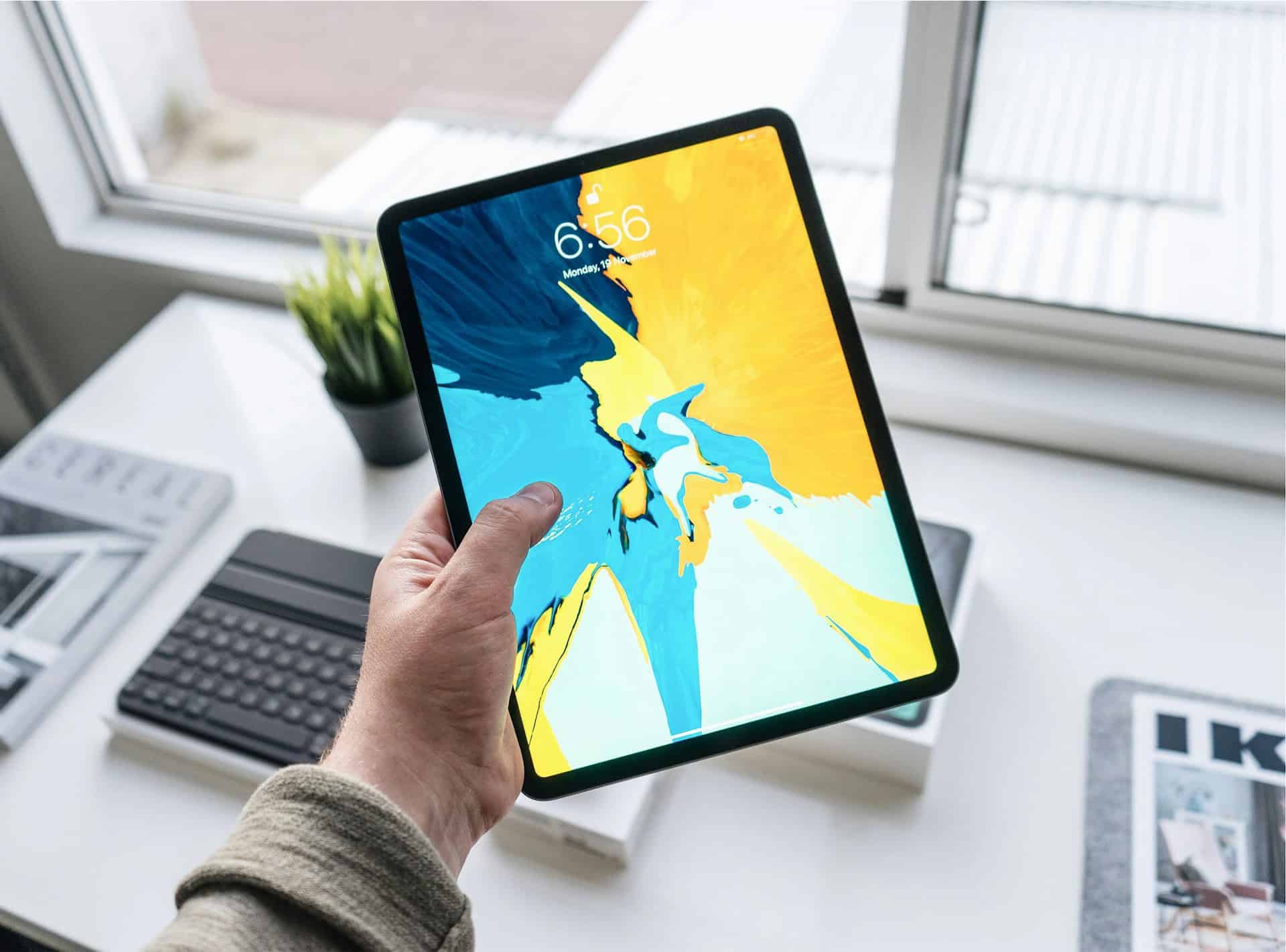 A person holding an iPad displaying a colorful abstract wallpaper, with a plant and keyboard visible on a desk in a well-lit room.