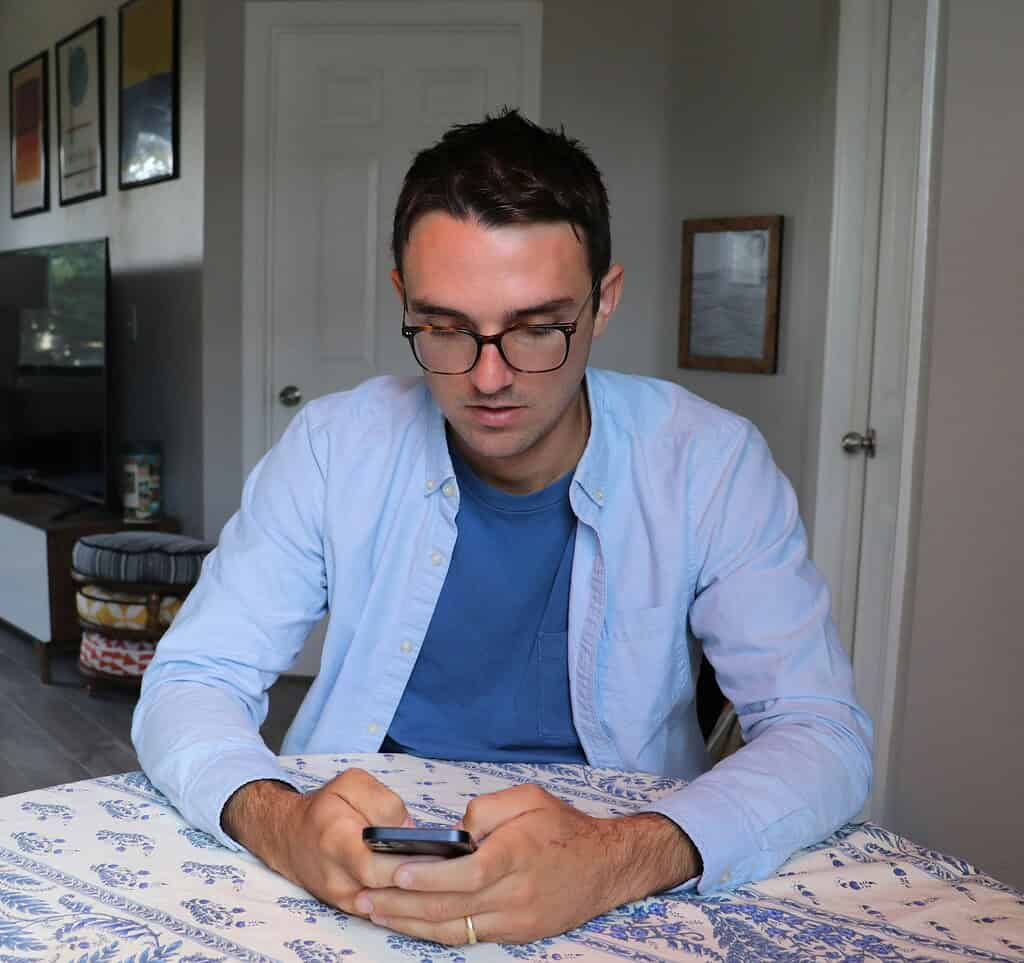 A seated man in a light blue shirt and blue t-shirt is focused on his smartphone, with a patterned tablecloth in a cozy room.