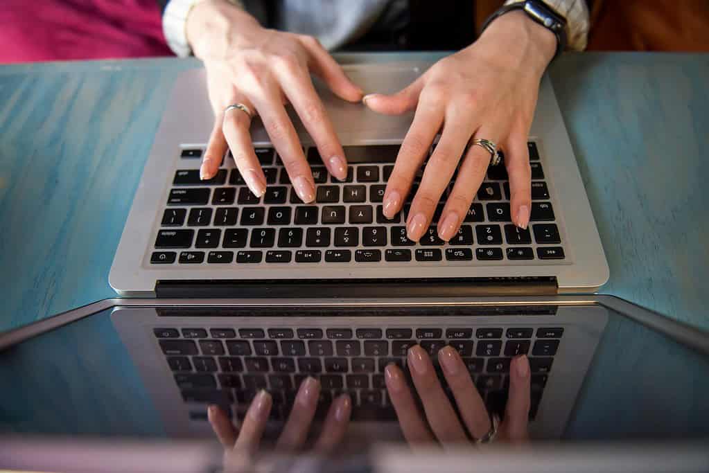 Overhead view of hands typing on a MacBook, reflected in a glossy blue desk surface, person wears rings and an Apple Watch.
