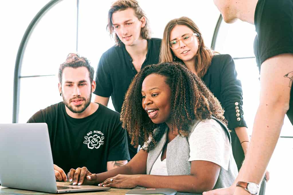 A diverse group of individuals collaborates around a laptop, focused on their work in a bright, modern workspace.