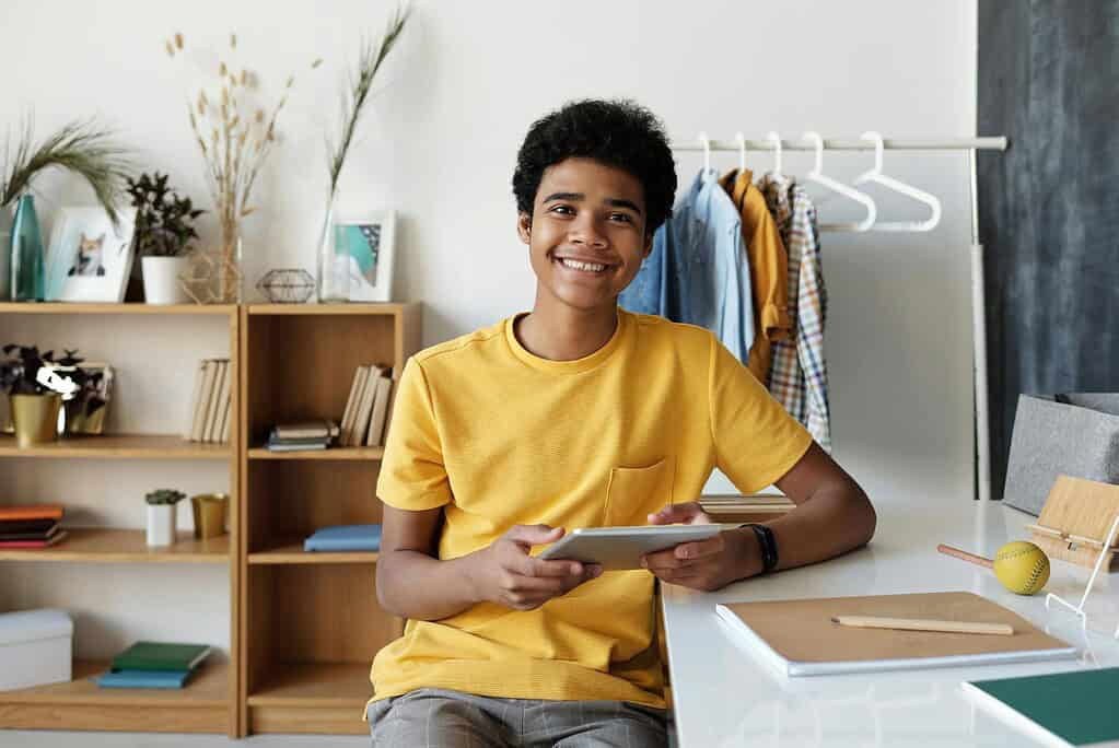 A young person in a yellow shirt sits at a desk holding a tablet, surrounded by books, plants, and colorful clothing in a bright room.