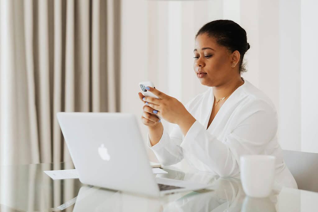 A woman in a white blouse sits at a glass table, focused on her smartphone, with a laptop and a white mug nearby.