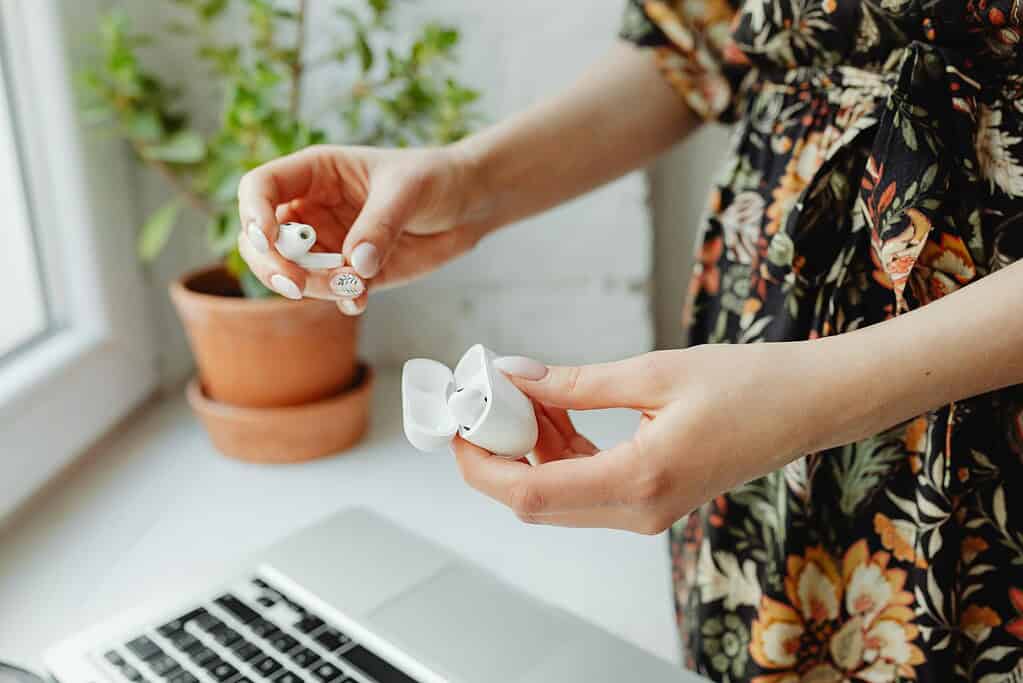 A person holds wireless earbuds near a charging case, with a laptop and potted plant visible in the background.