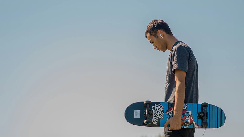 A person stands outdoors, holding a colorful skateboard with a graphic design, under a clear blue sky.
