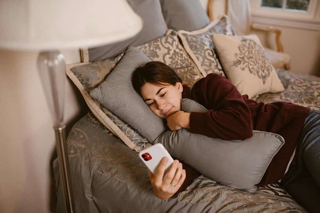 A person lying comfortably on a plush bed, holding a smartphone and relaxed against decorative pillows. Warm, soft lighting surrounds the scene.