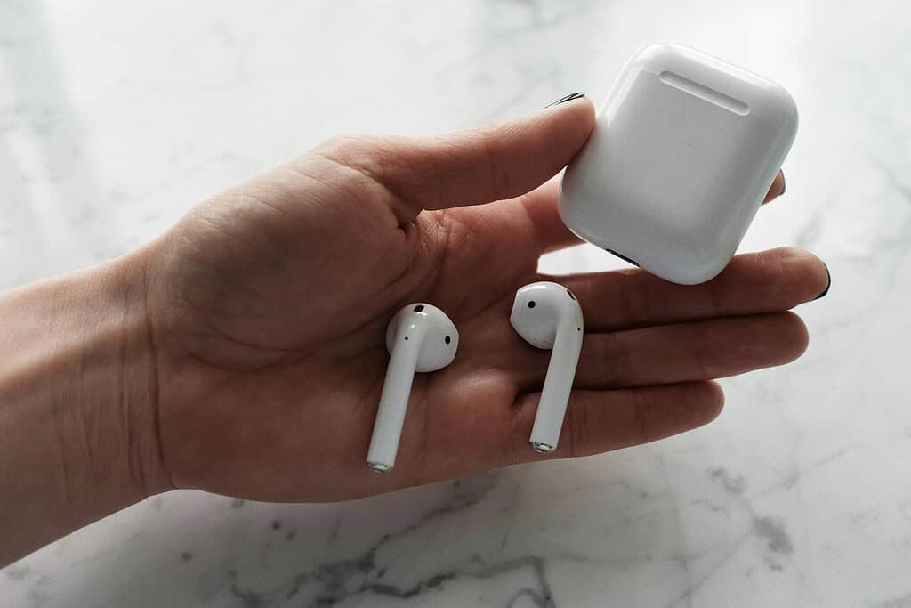 A hand holding two white wireless earbuds and their charging case on a marble surface.