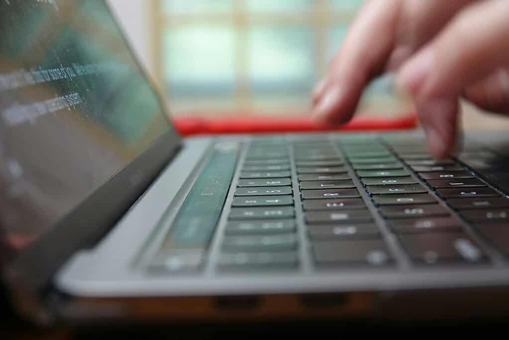 Close-up of fingers typing on a laptop keyboard, with a blurred screen displaying text in a well-lit indoor setting.