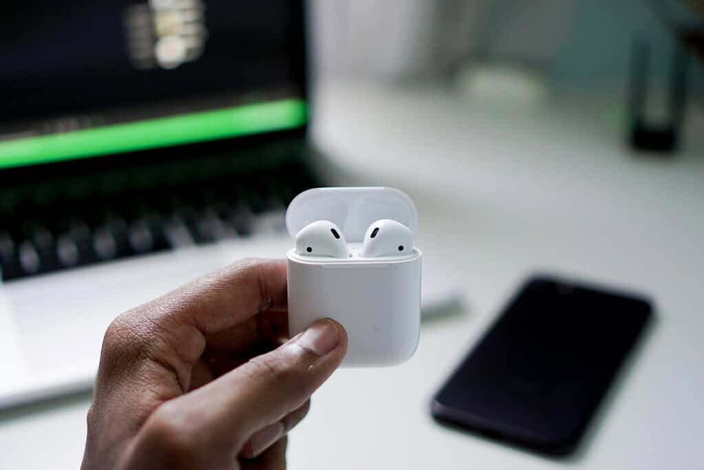 A hand holds a white AirPods case with earbuds inside, in front of a blurred laptop and smartphone on a white desk.