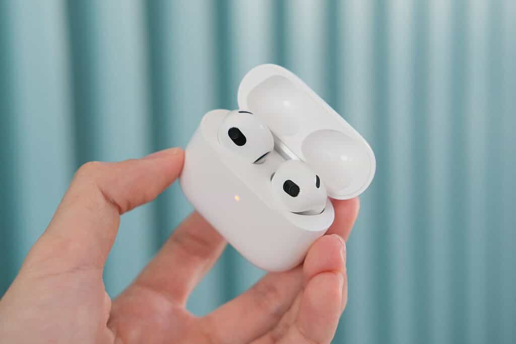 A hand holding an open white AirPods case, revealing two wireless earbuds inside, against a soft blue background.