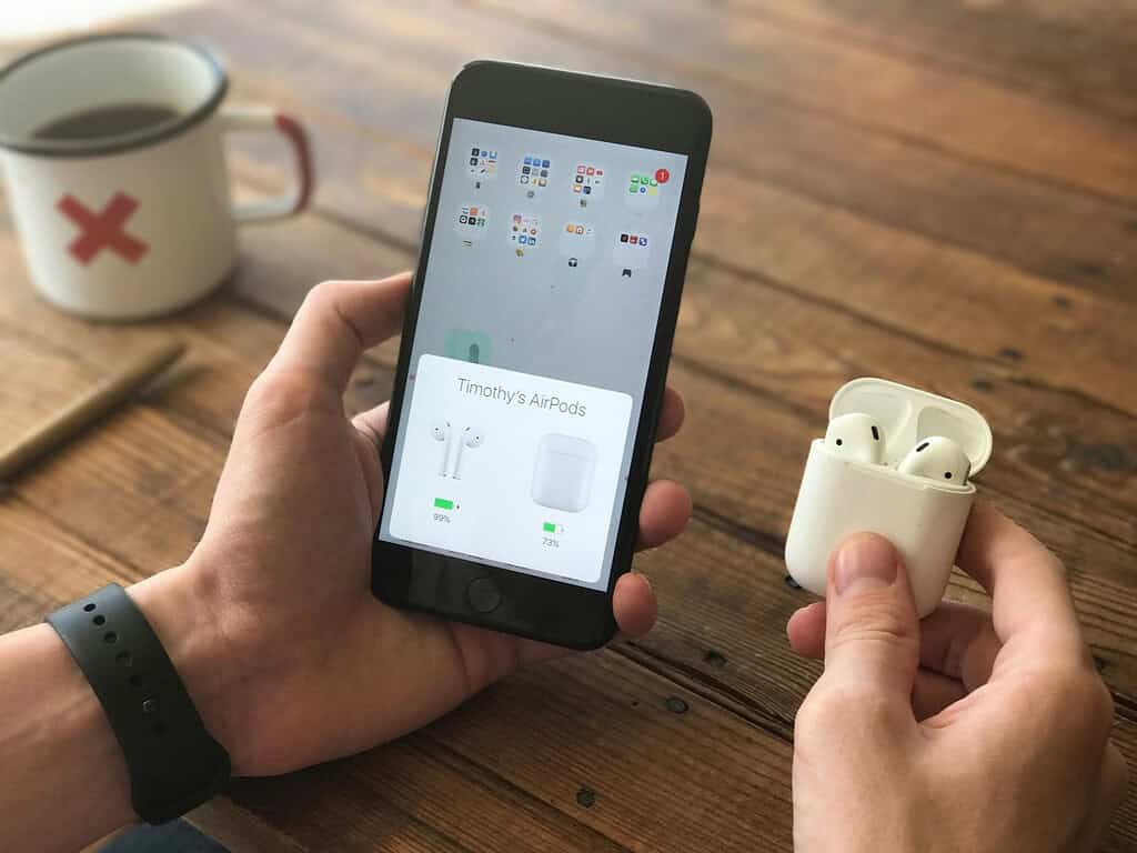 A hand holds an iPhone displaying AirPods battery levels, with white AirPods in their case nearby and a cup in the background.