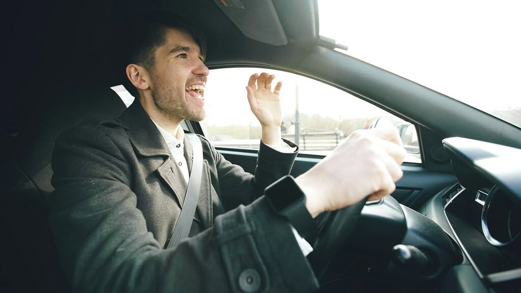A person wearing a dark coat drives a car, with one hand on the steering wheel and the other raised, set against a bright, sunny background.