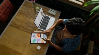 Person holding iPhone at a table with MacBook Pro showing code, iPad, Apple Pencil and AirPods