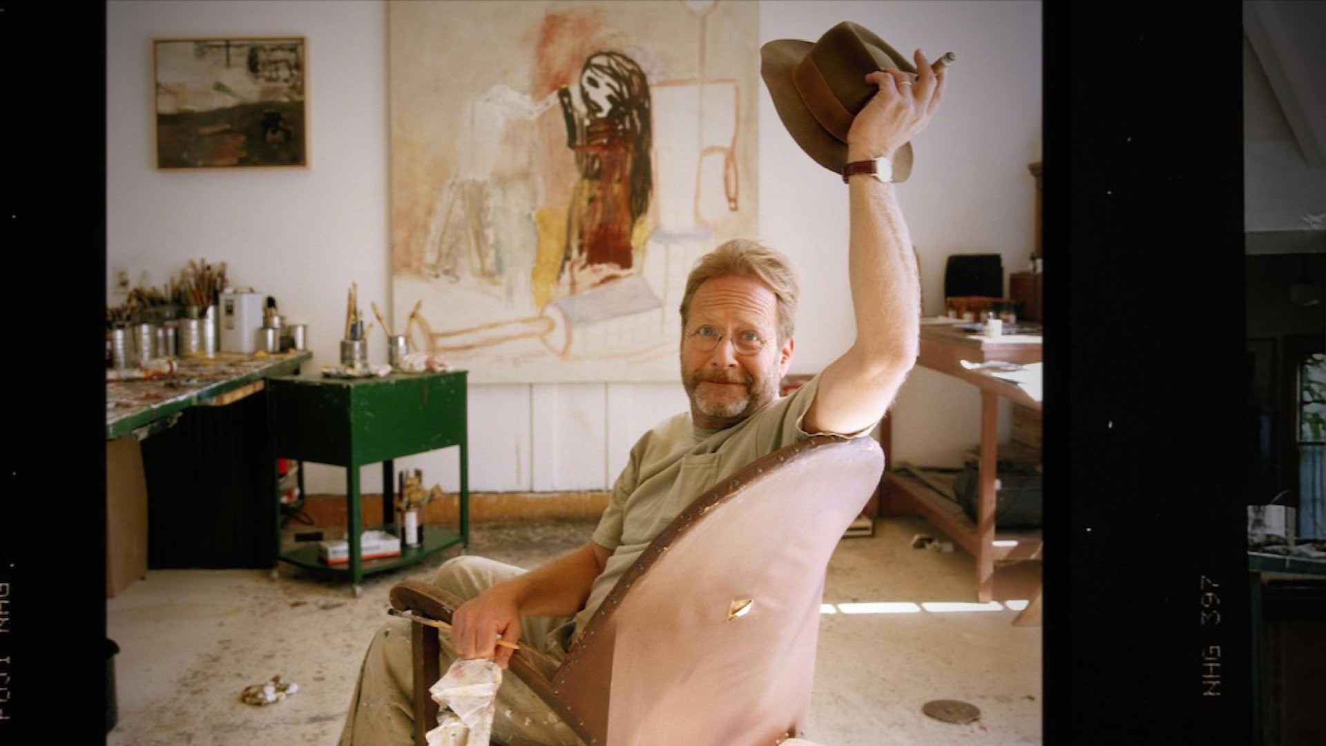 Martin Mull sitting in his art studio and raising his hat